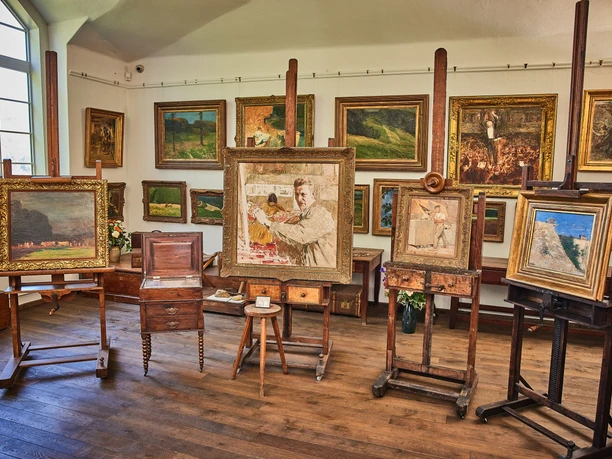 Een atelier met verschillende schildersezels waarop ingelijste schilderijen hangen; op de achtergrond hangen andere schilderijen aan de muur.