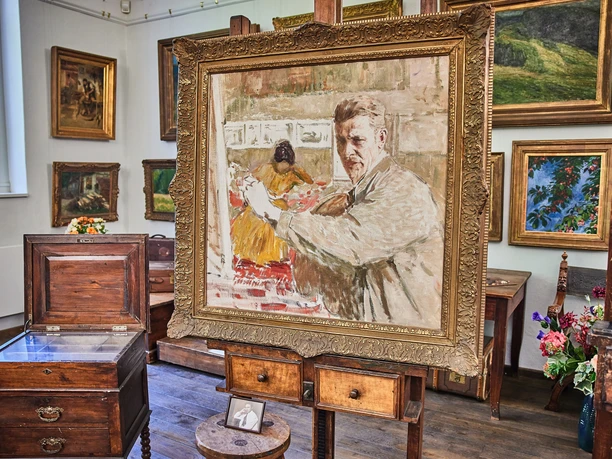 Robert Sterl Haus in Wehlen Ein Gemälde in einem goldenen Rahmen zeigt einen Mann beim Malen; umgeben von weiteren Gemälden und antiken Möbeln in einem hellen Raum.