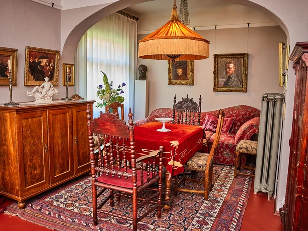 Robert Sterl Haus in Wehlen Gemütliches Wohnzimmer mit antiken Möbeln, rotem Teppich und Tischdecke, umgeben von Gemälden und einer großen, orangefarbenen Hängelampe.