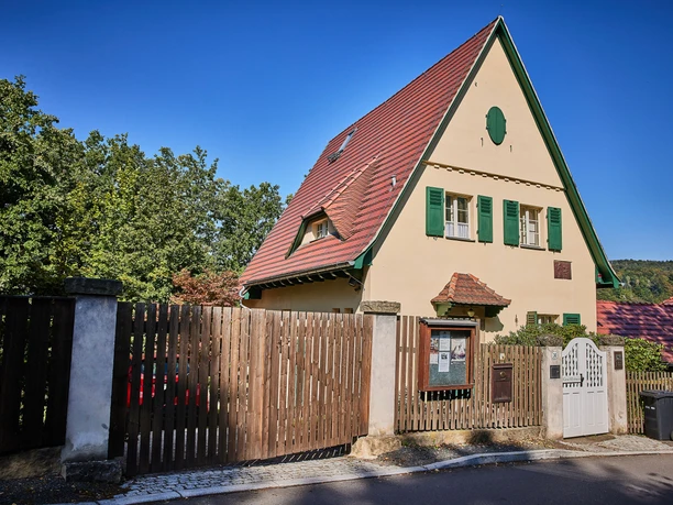 Een beige huis met een rood schuin dak en groene luiken, omringd door een houten hek, tegen een strakblauwe lucht.