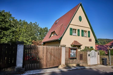 Robert Sterl Haus in Wehlen Ein beige Haus mit rotem Satteldach und grünen Fensterläden, umgeben von einem Holzzaun, vor einem klaren blauen Himmel.