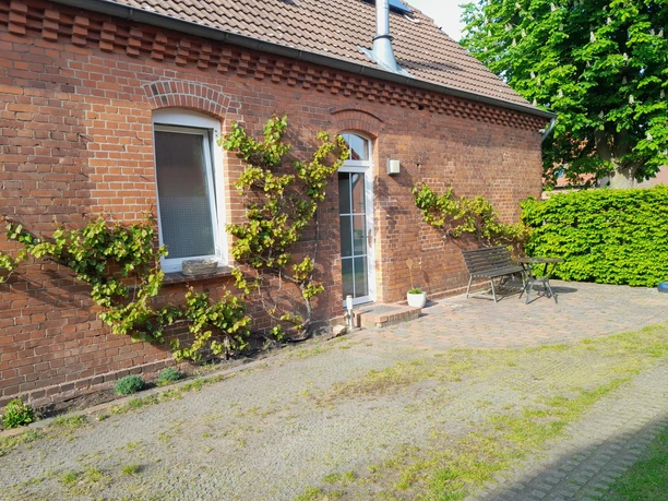 Dreppenstedt Rotes Backsteingebäude mit Rankpflanzen an der Fassade, davor Pflastersteinterrasse und Bank.