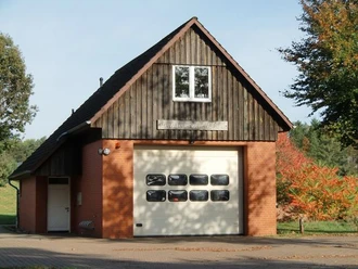 SpritzenhausWetzen.jpg Kleines Feuerwehrhaus aus rotem Backstein mit Holzgiebel, großer Torfront und Bäumen im Hintergrund.