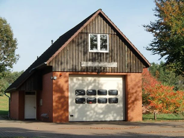 SpritzenhausWetzen.jpg Kleines Feuerwehrhaus aus rotem Backstein mit Holzgiebel, großer Torfront und Bäumen im Hintergrund.