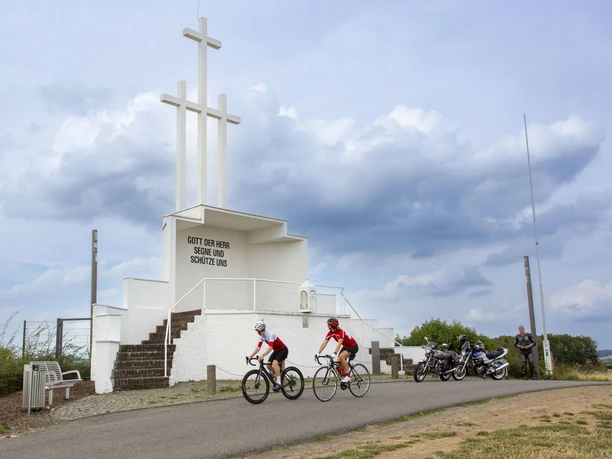 Ommerborner Kreuz Zwei Radfahrer fahren an einem weißen Denkmal mit drei Kreuzen bei leicht bewölktem Himmel vorbei.