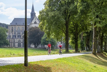 Altenberger Dom Radfahrer auf einem baumgesäumten Weg, mit einer historischen Kirche im Hintergrund bei sonnigem Wetter.