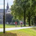 Altenberger Dom Radfahrer auf einem baumgesäumten Weg, mit einer historischen Kirche im Hintergrund bei sonnigem Wetter.
