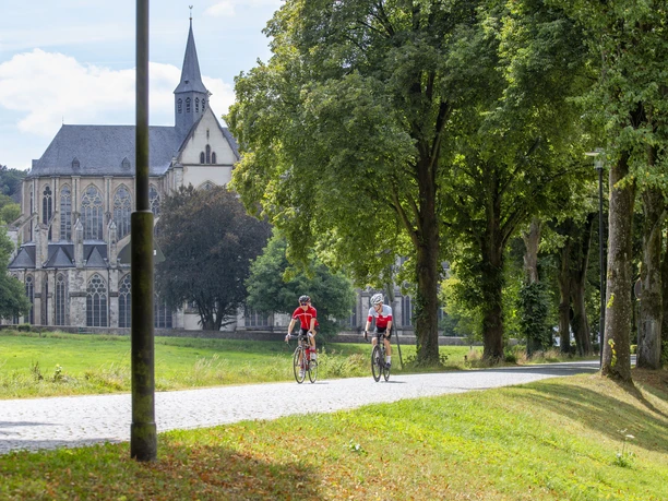 Altenberger Dom Radfahrer auf einem baumgesäumten Weg, mit einer historischen Kirche im Hintergrund bei sonnigem Wetter.
