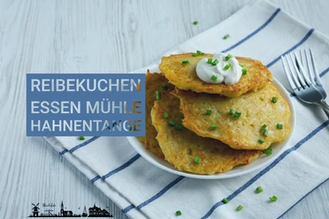 Himmelfahrt Mühle Hahnentange-1.jpg Drei goldbraune Reibekuchen auf einem weißen Teller mit einem Klecks Sauerrahm und Schnittlauch, daneben eine Gabel auf einem blau-weiß karierten Tuch.
