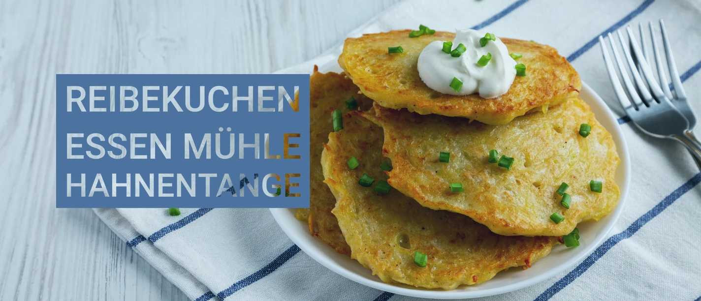 Himmelfahrt Mühle Hahnentange-1.jpg Drei goldbraune Reibekuchen auf einem weißen Teller mit einem Klecks Sauerrahm und Schnittlauch, daneben eine Gabel auf einem blau-weiß karierten Tuch.