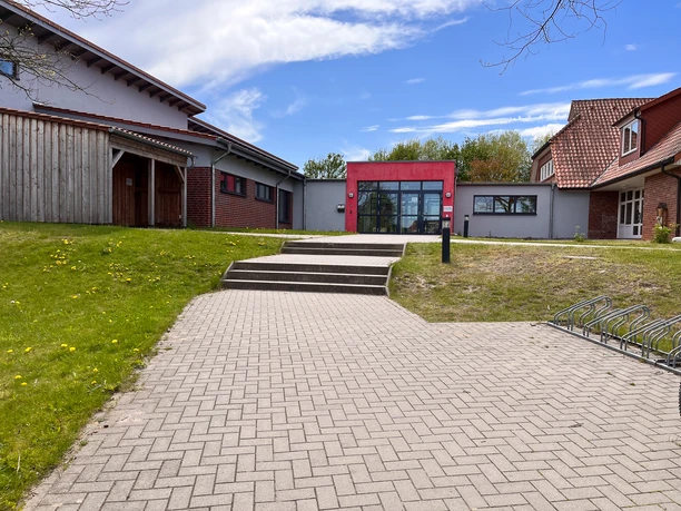 IMG_0760.jpg Außenansicht des Kindergartens mit rotem Eingangsbereich und gepflastertem Weg.