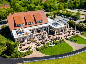 Luftaufnahme eines großen Gebäudes mit roten Ziegeldach, Sonnenterrasse und grünem Park, umgeben von Bäumen.