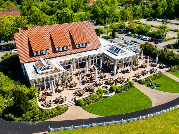 Halle-TerrasseRossiniDrohne-TeutoburgerWald-mayfeldWerbeagentur-HenningMayer.jpg Luftaufnahme eines großen Gebäudes mit roten Ziegeldach, Sonnenterrasse und grünem Park, umgeben von Bäumen.