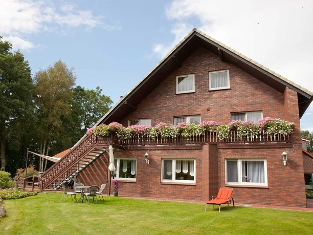 Bauerncafé.jpg Backsteingebäude mit Balkon und blühenden Geranien, umgeben von einer grünen Wiese.