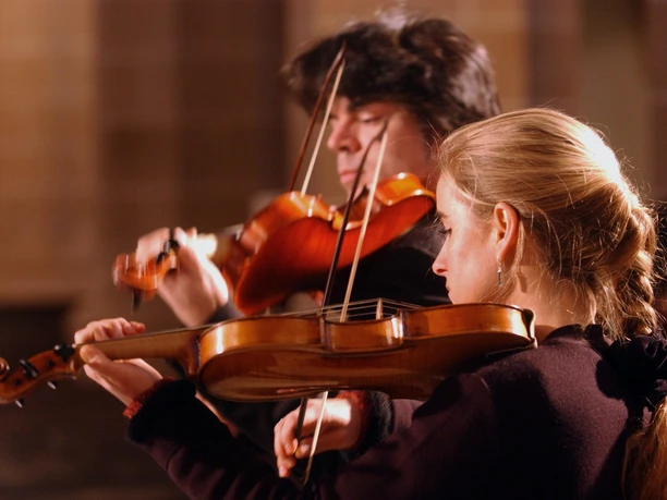 Mozart in Sachsen 1.jpg Zwei Musiker spielen Geigen in einem Konzertsaal, stimmungsvoll beleuchtet.