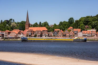 Blick auf Elbe und Lauenburg Ein Schiff fährt auf einem Fluss. Am Ufer steht eine Stadt mit einer Kirche.