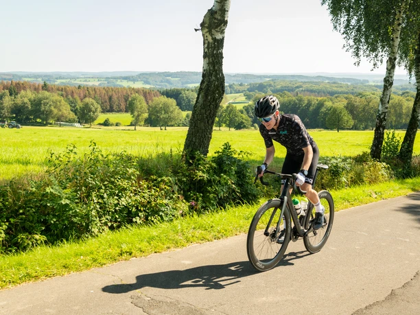 Radfahrer fährt auf einer ländlichen Straße, umgeben von grüner Landschaft und sanften Hügeln.