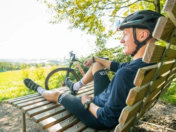 Pause am Heckberg Radfahrer sitzt entspannt auf einer rustikalen Holzbank mit Blick auf eine malerische, grüne Landschaft.