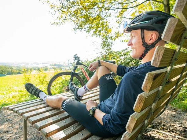 Radfahrer sitzt entspannt auf einer rustikalen Holzbank mit Blick auf eine malerische, grüne Landschaft.