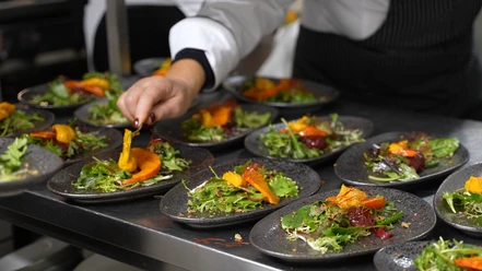Ein Koch garniert sorgfältig Teller mit frischem Salat und gebratenen Kürbisspalten in einer Küche.