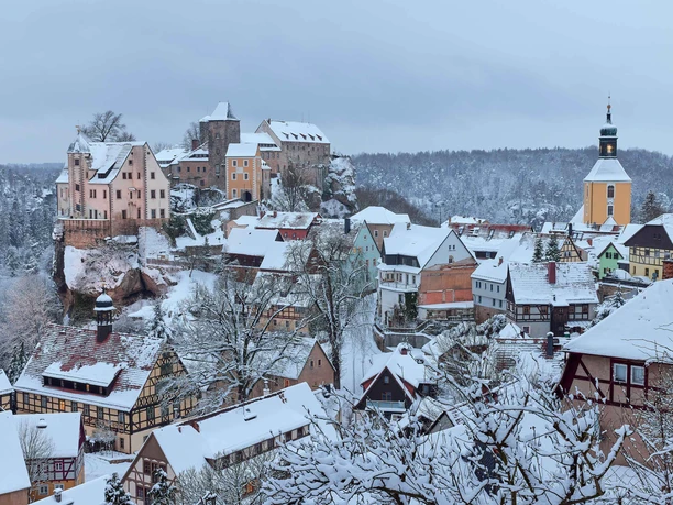 Snow-covered Hohnstein Castle towers over a wintry village in the Elbe Sandstone Mountains, surrounded by forests.