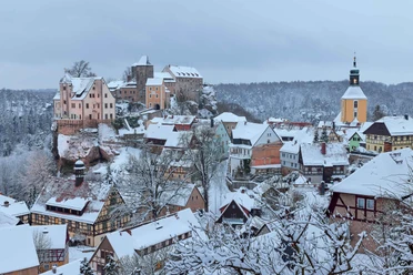 Burg Hohnstein Verschneite Burg Hohnstein thront über einem winterlichen Dorf im Elbsandsteingebirge, umgeben von Wäldern.