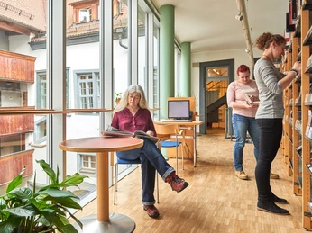 Lesen in der Stadtbibliothek in Pirna Drei Frauen in einer Bibliothek; eine sitzt am Tisch und liest, zwei stehen an Regalen. Helle, moderne Atmosphäre mit großen Fenstern.