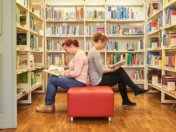 Two women sit back to back on a red stool in a library and read books; surrounded by full bookshelves.