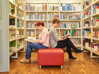 Lesen in der Stadtbibliothek Zwei Frauen sitzen Rücken an Rücken auf einem roten Hocker in einer Bibliothek und lesen Bücher; umgeben von vollen Bücherregalen.
