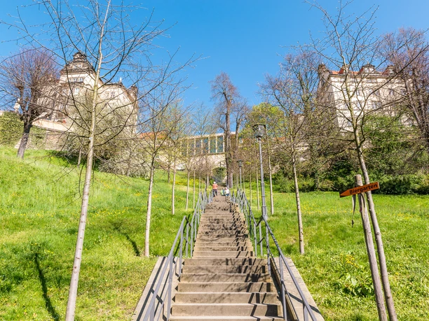 Stone steps lead through a green meadow to a castle surrounded by bare trees; a sign points to the beer garden.