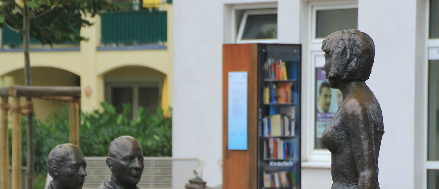 Skulpturengruppe mit Menschen aus Bronze, die rund um einen Tisch sitzen, im Freien positioniert.