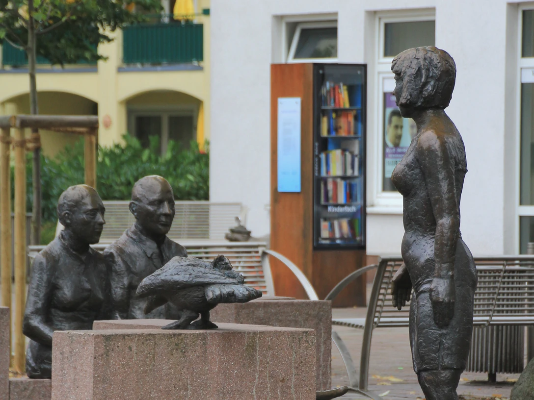Kunst_und_Buecher_©_DKiener.jpg Skulpturengruppe mit Menschen aus Bronze, die rund um einen Tisch sitzen, im Freien positioniert.