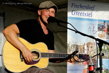 Sommer Konzerte Greetsiel: Oliver Jüchems Musiker spielt lächelnd Gitarre und singt bei einem Sommerkonzert in Greetsiel auf einer Open-Air-Bühne.