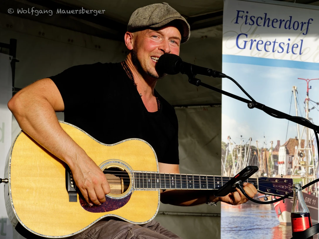 Sommer Konzerte Greetsiel: Oliver Jüchems Musiker spielt lächelnd Gitarre und singt bei einem Sommerkonzert in Greetsiel auf einer Open-Air-Bühne.
