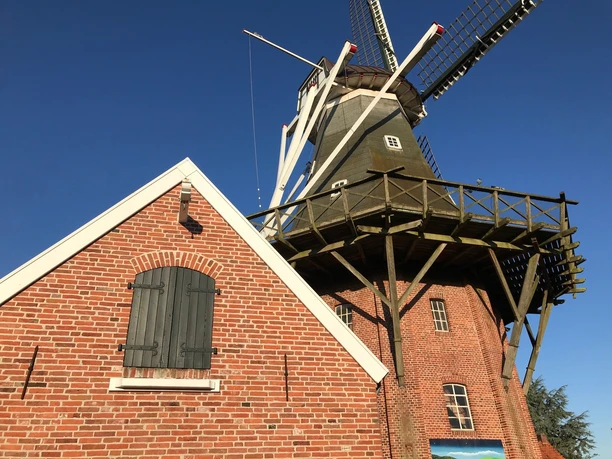 Mühle Rysum Historische Windmühle aus rotem Backstein mit Holzgalerie und weißen Flügeln unter blauem Himmel.