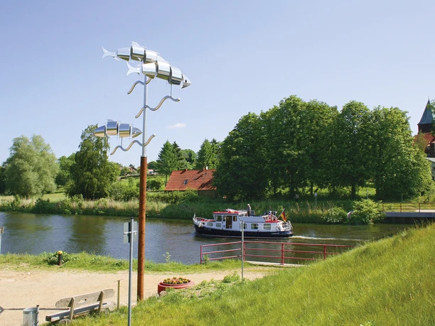 Kornbrennerweg Ein Schiff fährt auf dem Elbe-Lübeck-Kanal bei Krummesse. Am Ufer stehen grüne Bäume und eine Kirche. Die Sonne scheint.
