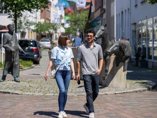 Halle-RonchinPlatz-Teutoburger-Wald-Tourismus-Patrick-Gawandtka-068_lowres.jpg Ein Paar spaziert bei Sonnenschein in einer Stadt, umgeben von modernen Skulpturen und Gebäuden.