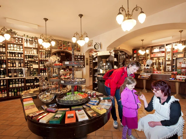Kaffeerösterei Schmole Ein gemütlicher Laden mit Regalen voller Weinflaschen; eine Frau in Schürze zeigt einem Kind in rosa Jacke etwas, während eine andere Frau zusieht.