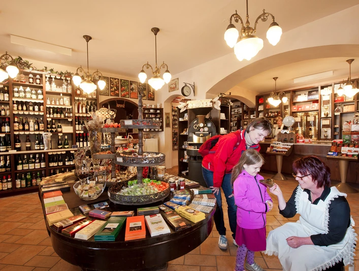 Schmole coffee roastery Ein gemütlicher Laden mit Regalen voller Weinflaschen; eine Frau in Schürze zeigt einem Kind in rosa Jacke etwas, während eine andere Frau zusieht.A cozy store with shelves full of bottles of wine; a woman in an apron shows a child in a pink jacket something while another woman looks on.Útulný obchůdek s regály plnými lahví vína; žena v zástěře ukazuje dítěti v růžové bundě, zatímco jiná žena se dívá.Przytulny sklep z półkami pełnymi butelek wina; kobieta w fartuchu pokazuje coś dziecku w różowej kurtce, podczas gdy inna kobieta patrzy.Een gezellige winkel met schappen vol flessen wijn; een vrouw in een schort laat een kind in een roze jasje iets zien terwijl een andere vrouw toekijkt.Un negozio accogliente con scaffali pieni di bottiglie di vino; una donna con un grembiule mostra qualcosa a un bambino con una giacca rosa mentre un'altra donna guarda.