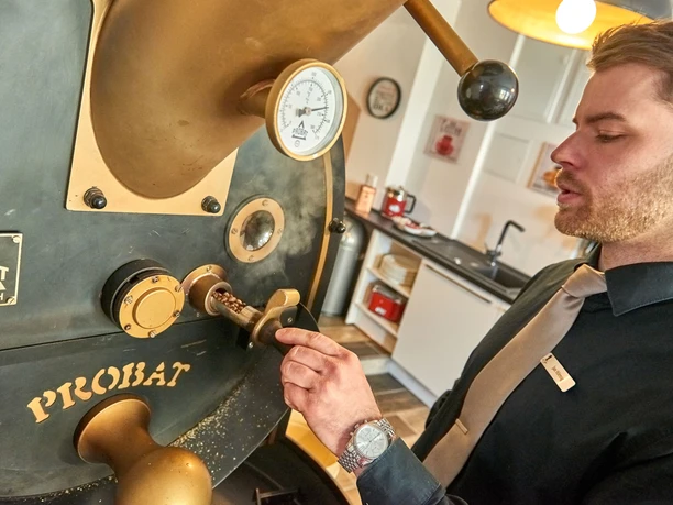 A man in a black shirt and tie operates a large, black coffee roasting machine with golden accents in a modern kitchen.