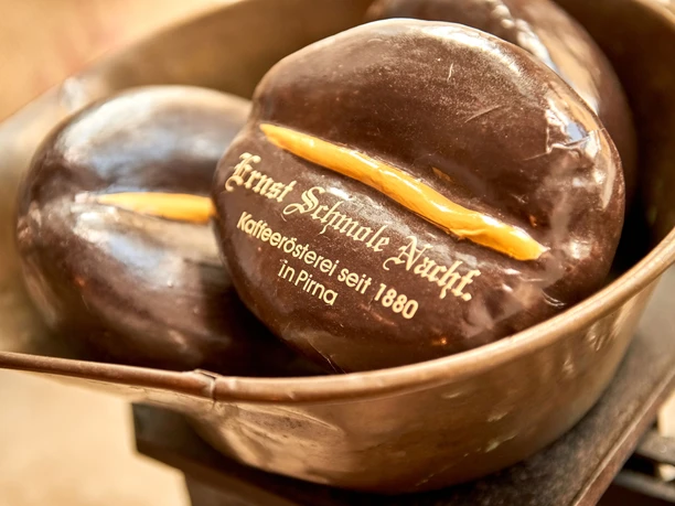 Twee grote, glanzende chocolade koffiebonen met een gele streep en de inscriptie "Ernst Schmole Nachf. Kaffeerösterei seit 1880 in Pirna" in een metalen kom.