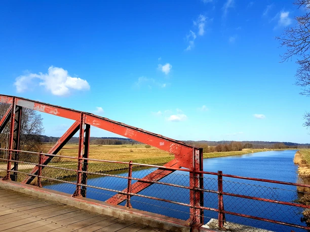 Eine rote Brücke führt über einen Fluss. Die Sonne scheint und der Himmel ist blau.