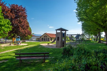 Reelkirchen_Spielplatz_2462_RHa.jpg Spielplatz mit Holzspielgeräten, umgeben von Bäumen und Bänken, bei sonnigem Wetter in einer grünen Umgebung.
