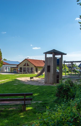 Reelkirchen_Spielplatz_2462_RHa.jpg Spielplatz mit Holzspielgeräten, umgeben von Bäumen und Bänken, bei sonnigem Wetter in einer grünen Umgebung.