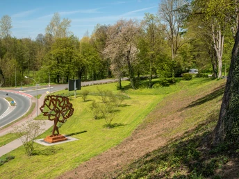 Skulptur eines Baumes auf einer Wiese neben Straßenkreuzung, umgeben von Bäumen und frischem Grün.