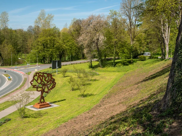 Nelkenpark_2403_RHa.jpg Skulptur eines Baumes auf einer Wiese neben Straßenkreuzung, umgeben von Bäumen und frischem Grün.