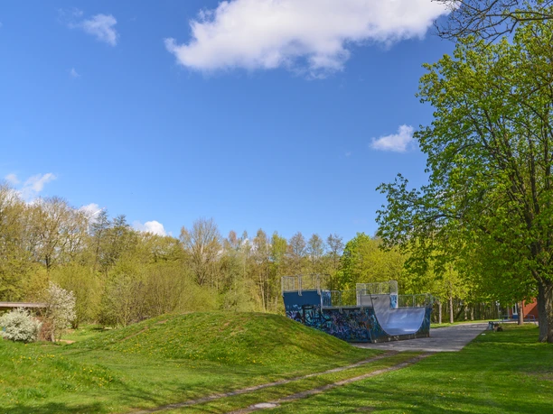Blomberg_JUZ_Scaterbahn_2216_RHa.jpg Skaterbahn in grüner Parksze, umgeben von Bäumen, mit einer Halfpipe und blauem Himmel im Hintergrund.