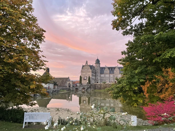 Schloss Hämelschenburg_Herbst 2022_Teichansicht_© Schloss Hämelschenburg.jpeg Schloss Hämelschenburg spiegelt sich im Teich, umrahmt von herbstlich gefärbten Bäumen bei Sonnenuntergang.
