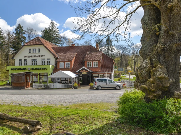 Blomberg_Forsthaus_2221_RHa.jpg Forsthaus Blomberg mit rustikalem Fachwerk und grünem Schild, umgeben von Bäumen, ein weißes Auto davor.