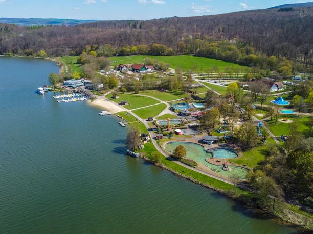 Schieder_Kronenbruch_0395_RHa.jpg Luftansicht eines Freizeitparks am Ufer eines Sees mit grünen Wiesen und bewaldeten Hügeln.